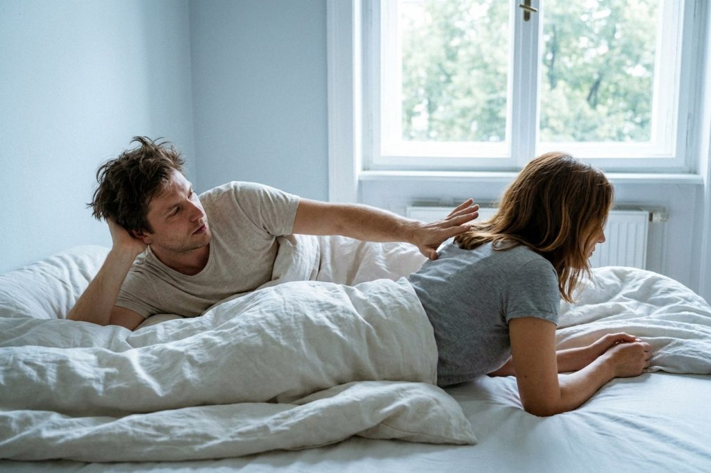 Im Bett streckt ein Mann die Hand nach der Schulter seiner Partnerin aus, die abgewandt von ihm liegt